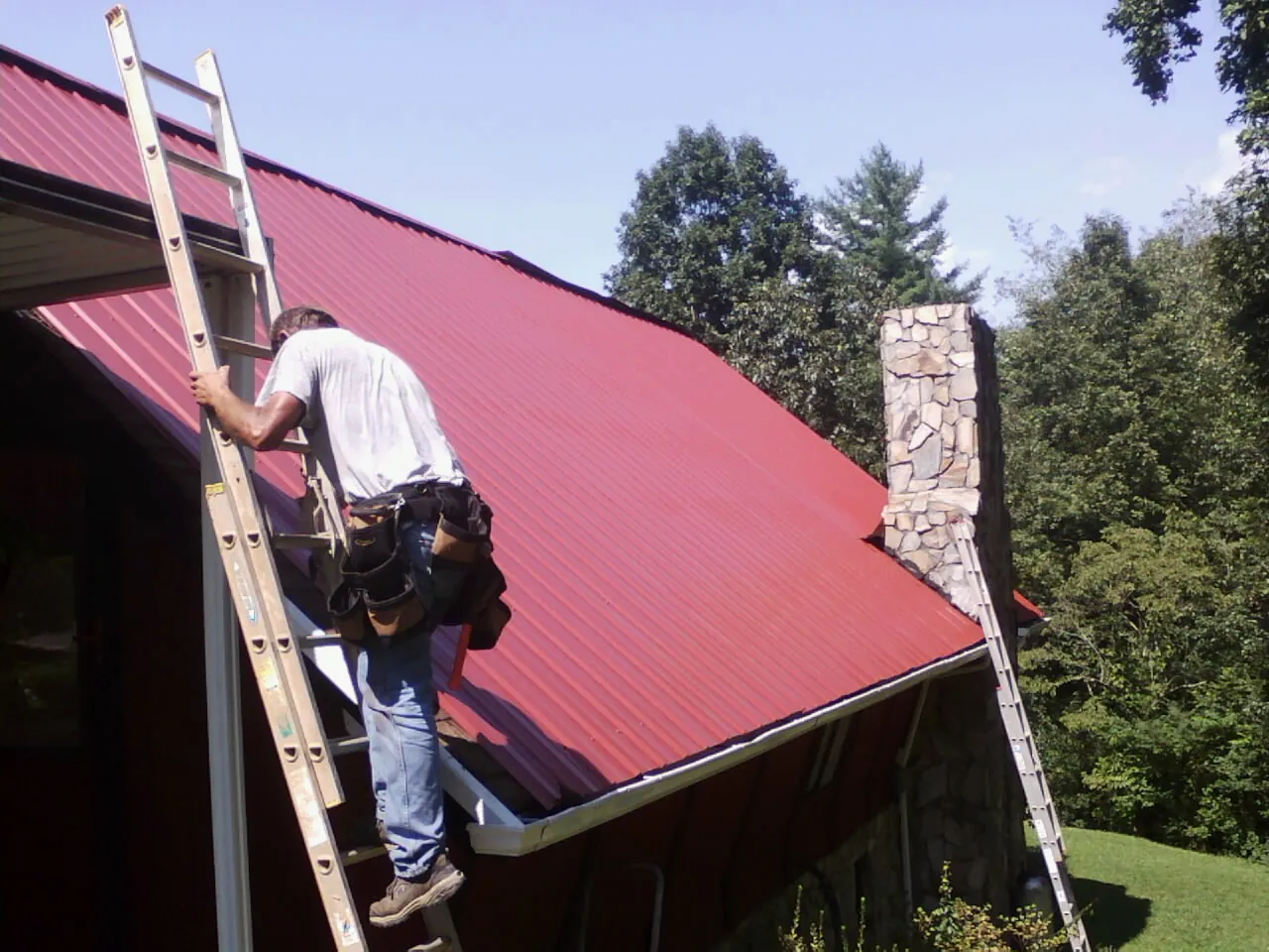 Roofer on a ladder beside a red metal roof and stone chimney — Soffit & Fascia Repair by Roofing Brothers in Depoe Bay